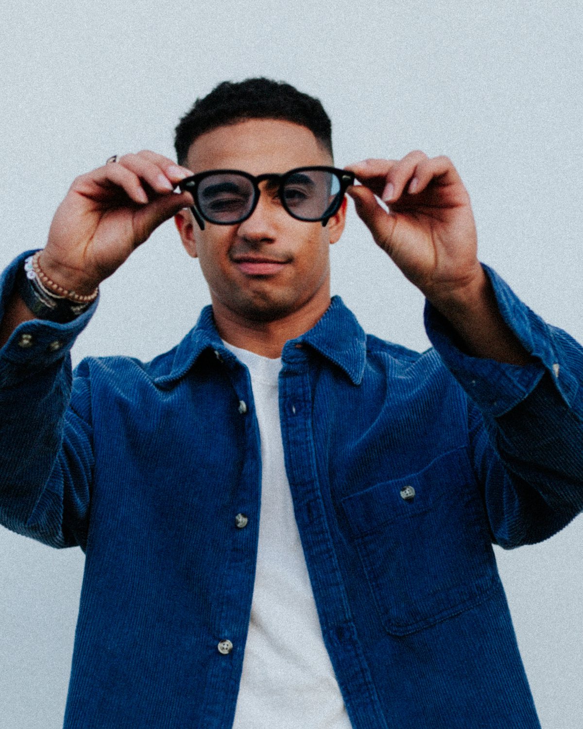 Cameron in a blue corduroy shirt, holding up a pair of glasses and squinting through them